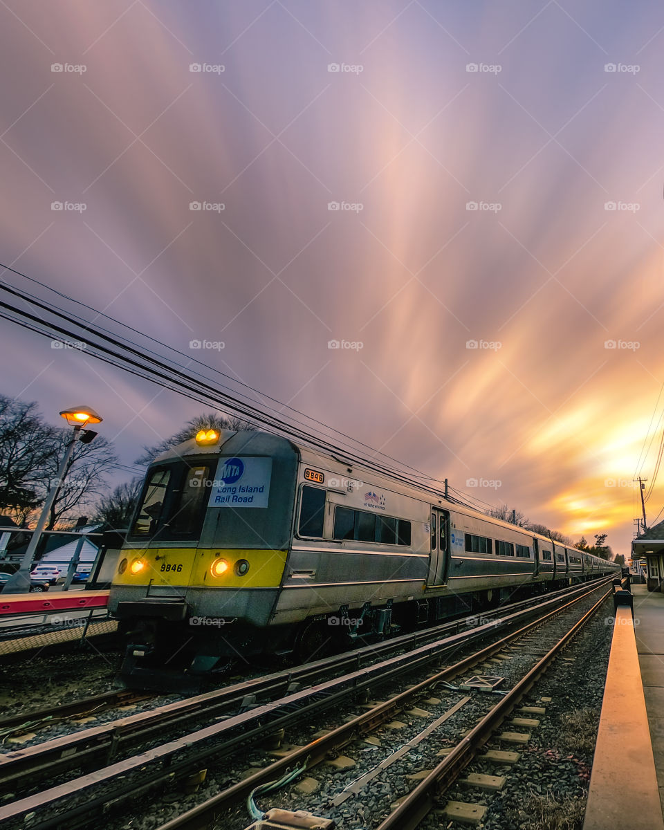 Long exposure of a commuter train sitting at a station as the clouds lit up by a setting sun streak across the sky. 