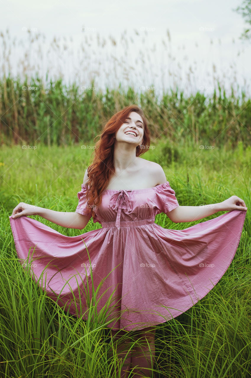 Beautiful portrait of young caucasian redhead woman dancing in the green field in pink dress.