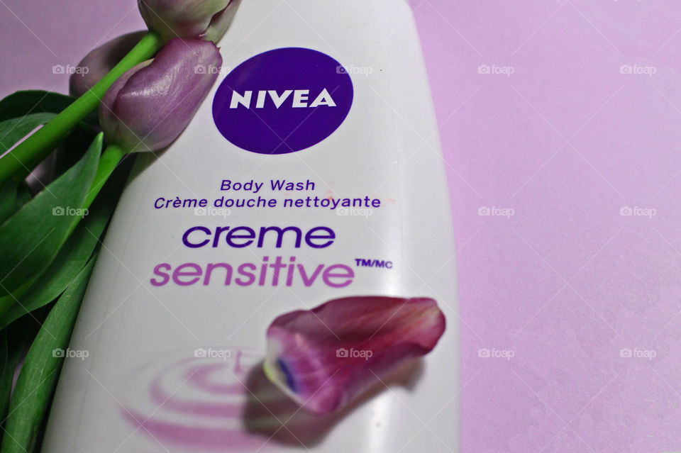 A flat-lay closeup of a bottle of NIVEA creme sensitive Body Wash with purple tulips on a pale violet background. One delicate purple and blue tinged petal rests on the bottle.
