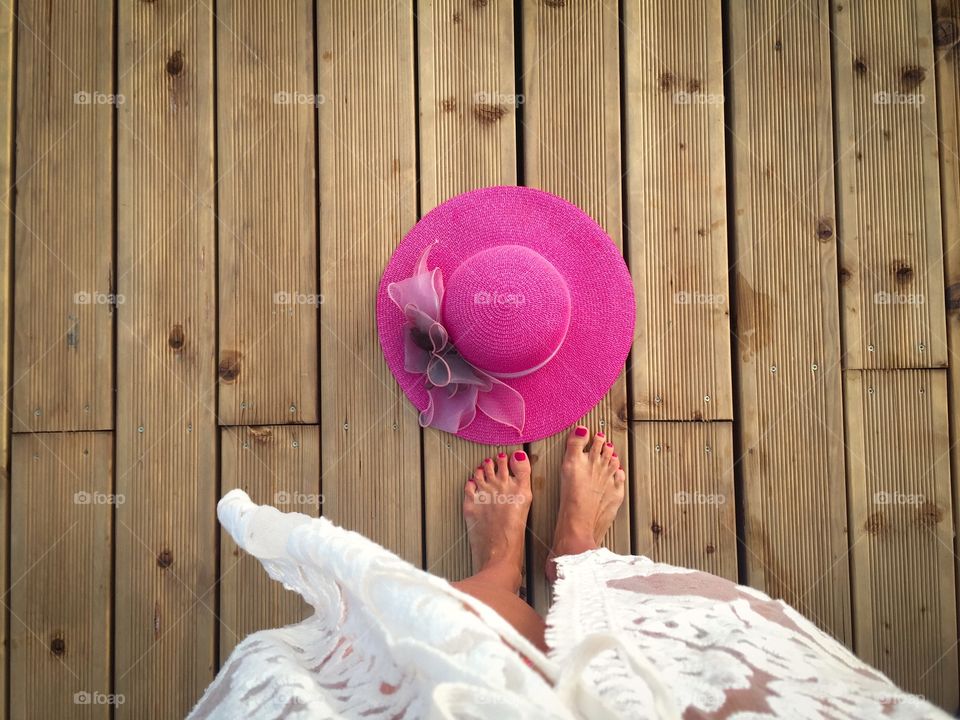 Looking down at woman wearing white dress standing near a pink summer hat on wooden pontoon