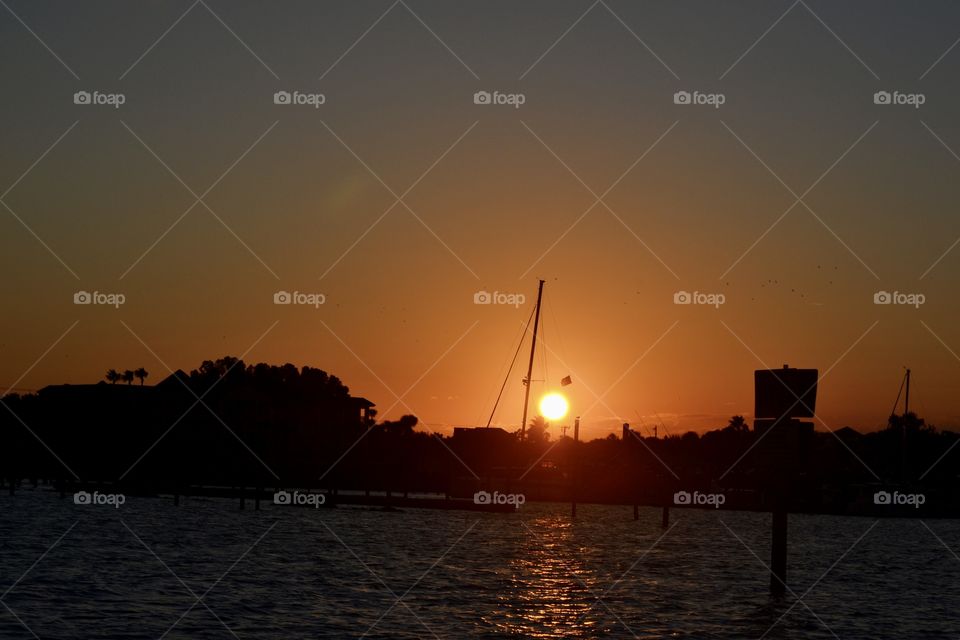A bright orange sun setting behind a sailboat on a river