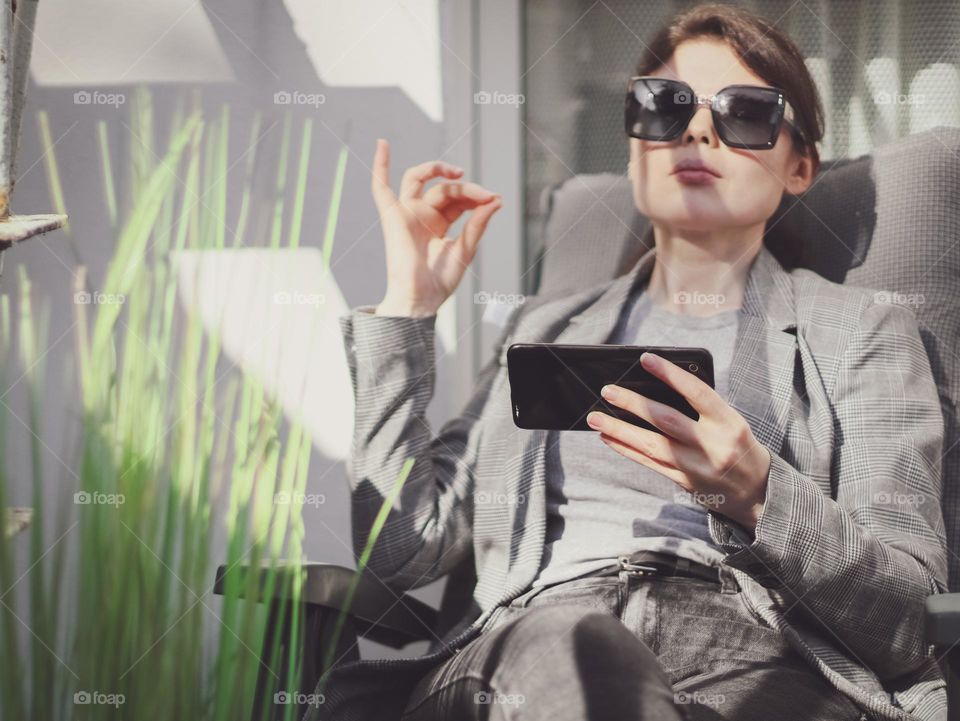 Portrait of a beautiful young Caucasian brunette girl in a gray jacket and a mobile phone in her hands sits meditating on a garden chair in the backyard of her house on a summer day, close-up side view. The concept of using technology, online meditat