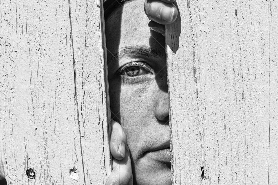 Women looking through a wooden fence. Women female is sad and trapped. Teardrop is running down the face of the women. The sunshine is shining on the face of the women and making shadows. 