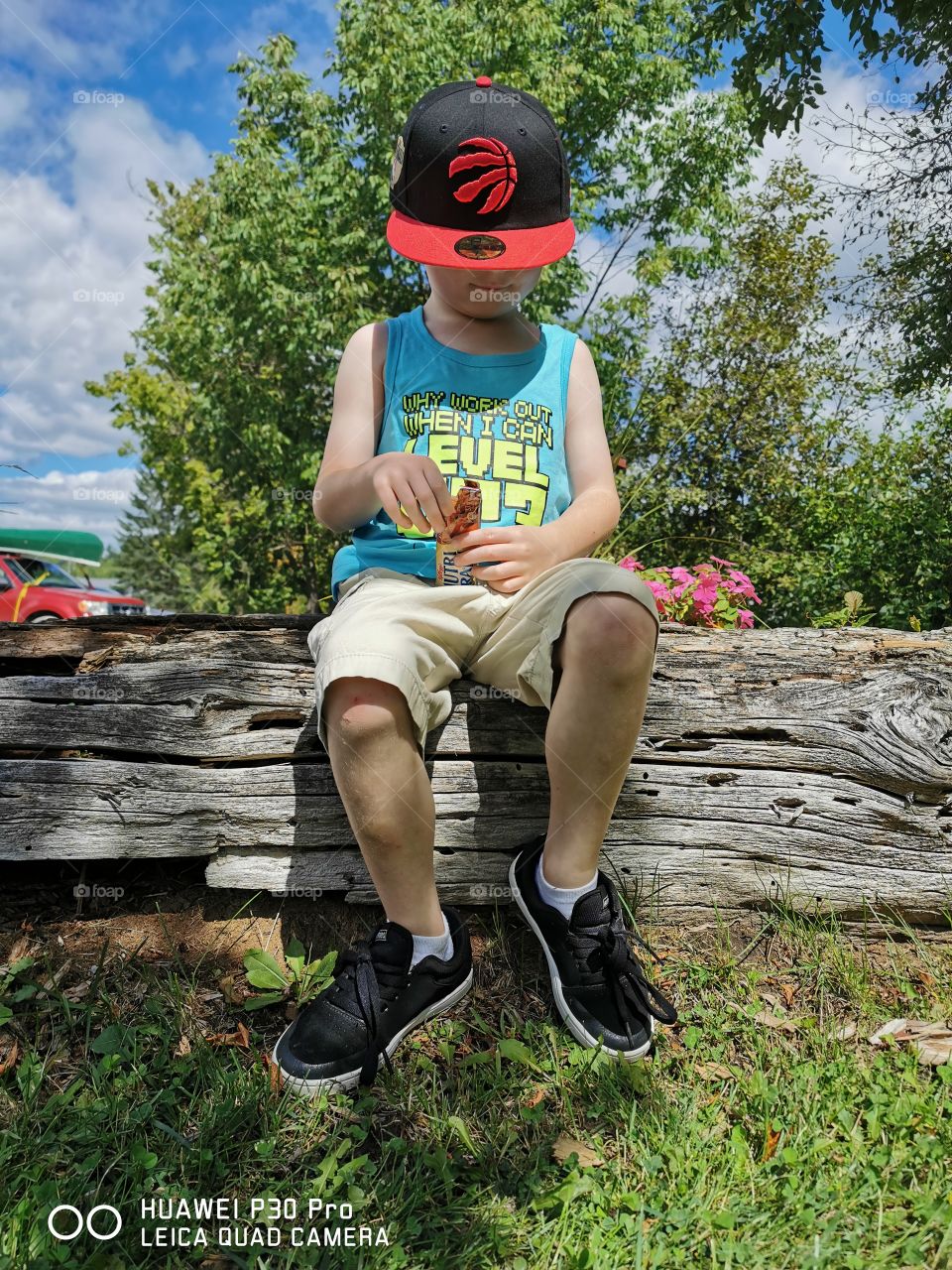 Young boy (my son) sitting on a log at a park, having a snack. Face covered by Raptors hat.