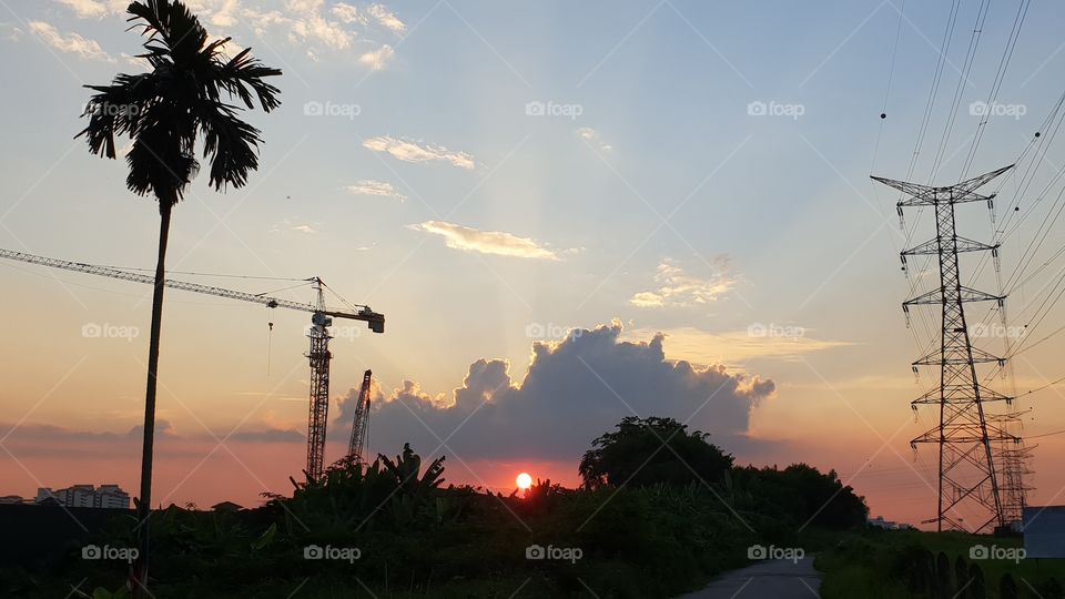 Sunset with the full Sun in view and clouds above, nestled between a tree and a signal tower. A contrasting symbol of nature and manmade.