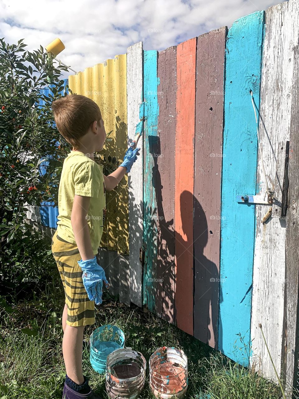 A child boy paints a fence with bright paint in the summer in the village, a funny shadow