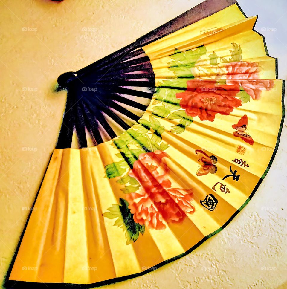 An Asian decorative fan, displayed on the wall decorated with red and green, flowers, leaves, butterflies, and Asian handwriting.