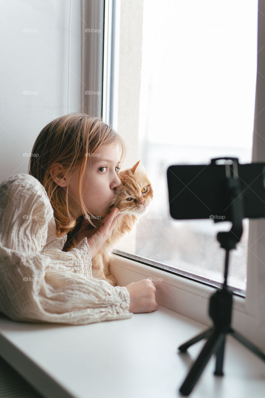 cute girl with her cat, sitting on the window and video chatting with friends. homemade red fluffy cat. Domestic pet
