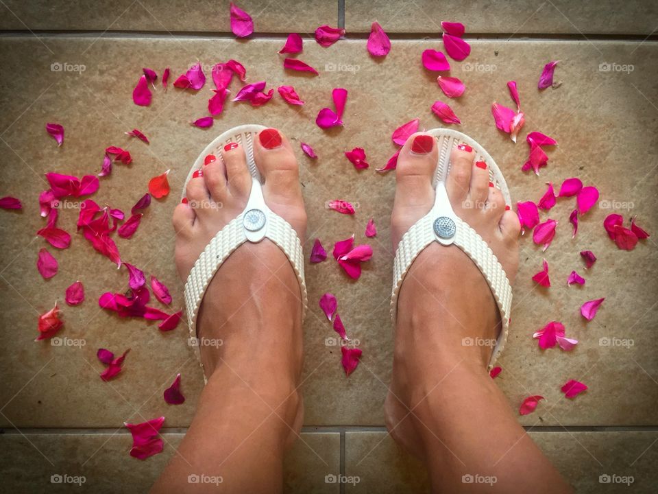 Looking down at woman’s feet wearing white flip flops surrounded by pink and red petals