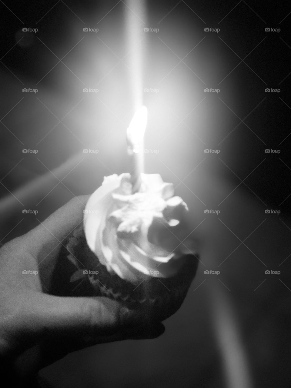 Black and white photo of a woman holding a chocolate cupcake with vanilla frosting and a glowing bright candle on top. 