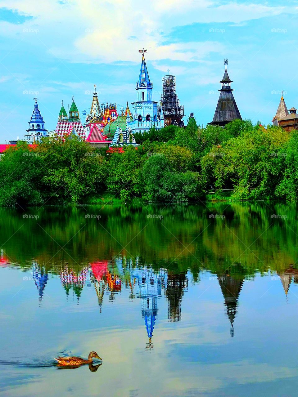 The domes of the towers and green trees are reflected in the water.  A duck is swimming in the foreground