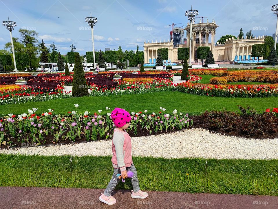 Nature. Plants. A little girl is walking in the park, she has a toy in her hands. In the background, colorful flowers beds