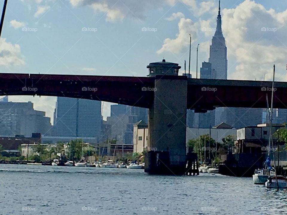The “Empire State Bldg” is visible behind the Long Island City skyline on the other side of the “Pulaski Bridge”, the large metallic structure spanning the East River. Various boats are lined up by the shore. 2018. Hypnotic Productions