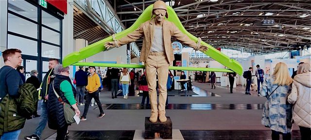 Is it a contraption for a circus act, some kind of projectile for a flying man or simply an oversized green clothes hanger he is attached to ? His goggles brought on the flight idea. It was seen at a trade show in “Munich”. 2024. Hypnotic Productions