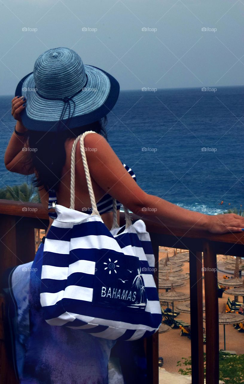 The girl in a hat and swimsuit looks into the distance to the sea. Striped beach bag on the shoulder