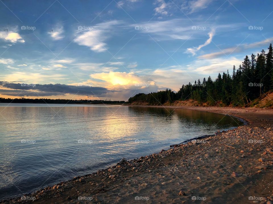 Sunset along a Canadian beach. Calm waters with spruce tree line. Rocky beach with nice clouds in sky. 