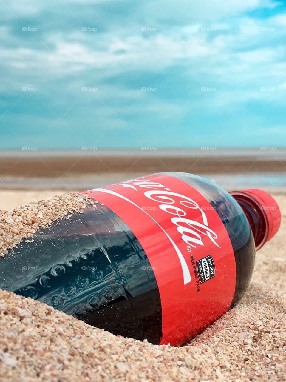 Bottle of unopened Coca Cola resting in sand on tropical beach suggesting a beach goer has it by his or her side. 