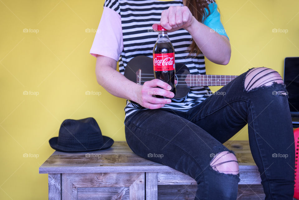 Young woman sitting on a desk with a ukulele and opening a bottle of Coca-cola