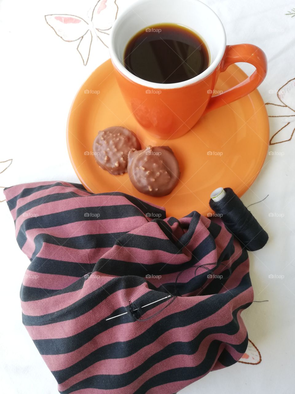 On a white patterned table cloth is an orange coffee mug on a plate with two chocolate biscuits. Next to them is a black and brown striped cloth. The hole will be sewn together with a needle and yarn.