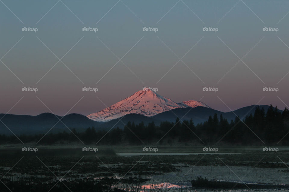 Mount Shasta catches on fire with the early morning sun rays, turning the mountain pink. The fog lifts from a nearby lake, giving an eery and mysterious feel to the mammoth and snow capped mountain. 