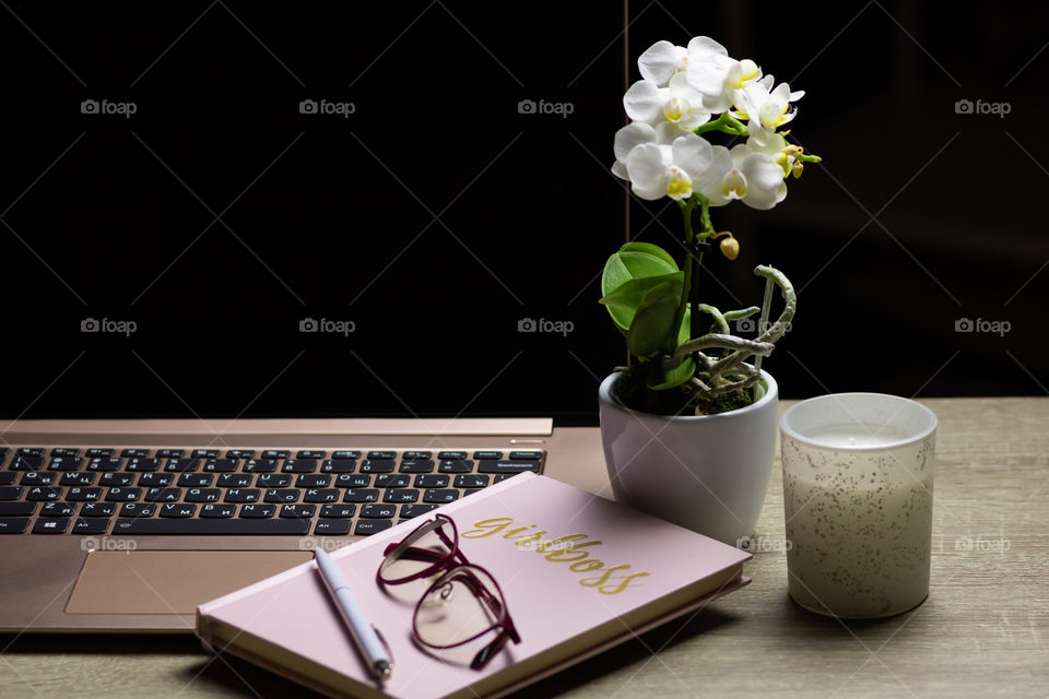 Still life in style workplace with computer, notebook, pen, glasses, orchid flower and candle on the table in the soft light.