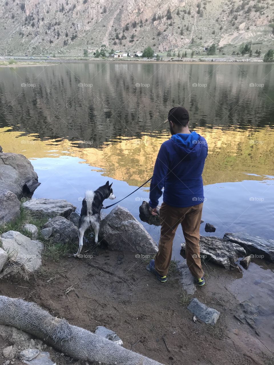 Man walks his Siberian Husky by a reflective mountain lake on a cool morning. This is an extremely casual shot.
