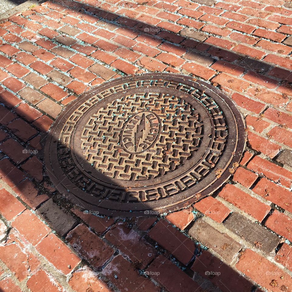 A manhole covers a power junction set in a brick walkway. Long shadows slant across them both.