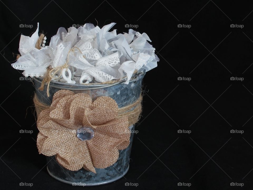An old fashioned brushed metal bucket with a burlap bow and white ribbon trinkets inside to the left of the frame with a solid black background with lots of copy space on the right. 