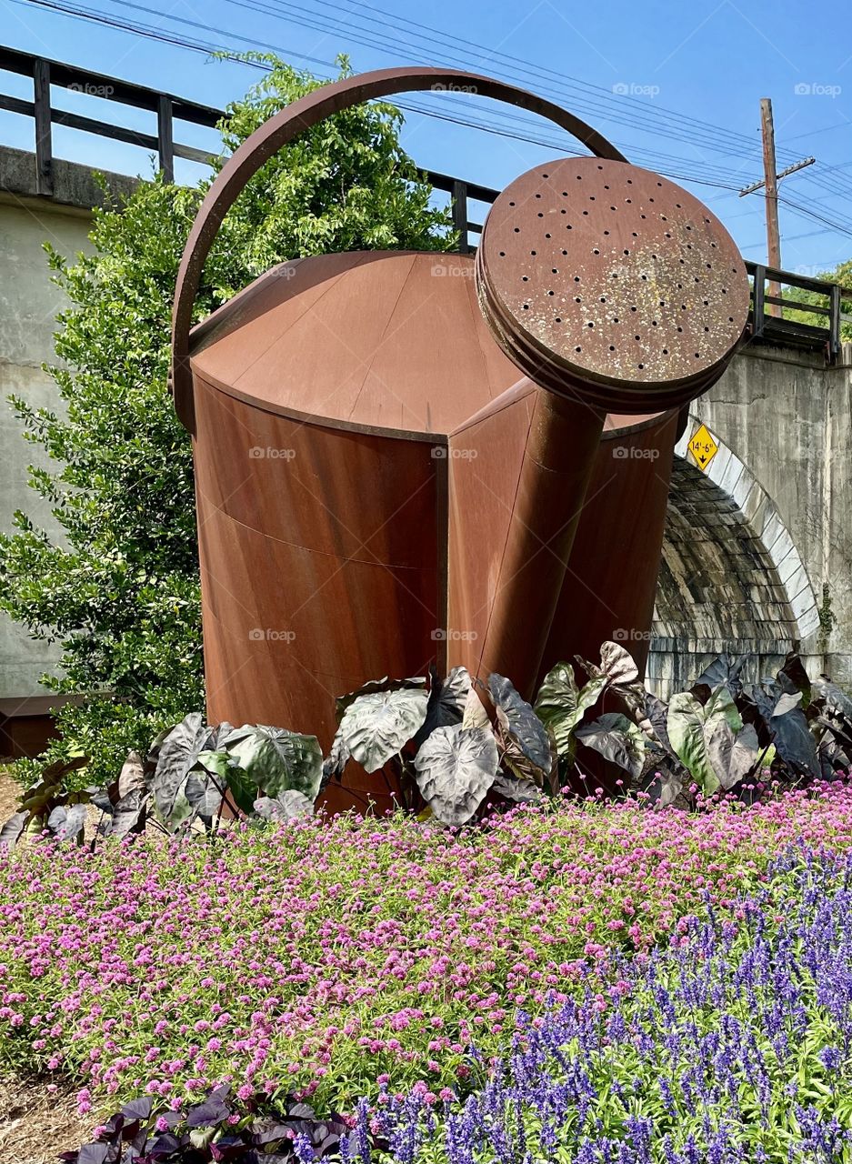 elephant ears, purple and pink annuals adorn the base of a watering can sculpture below a railroad tracks bridge in the city of Staunton,  Virginia 