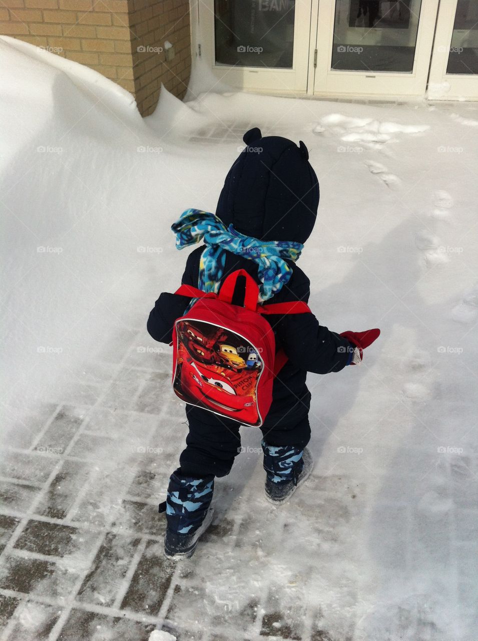 Toddler boy walks to school doors in snow while carrying a Disney Cars backpack and wearing a winter scarf, snowsuit and boots.