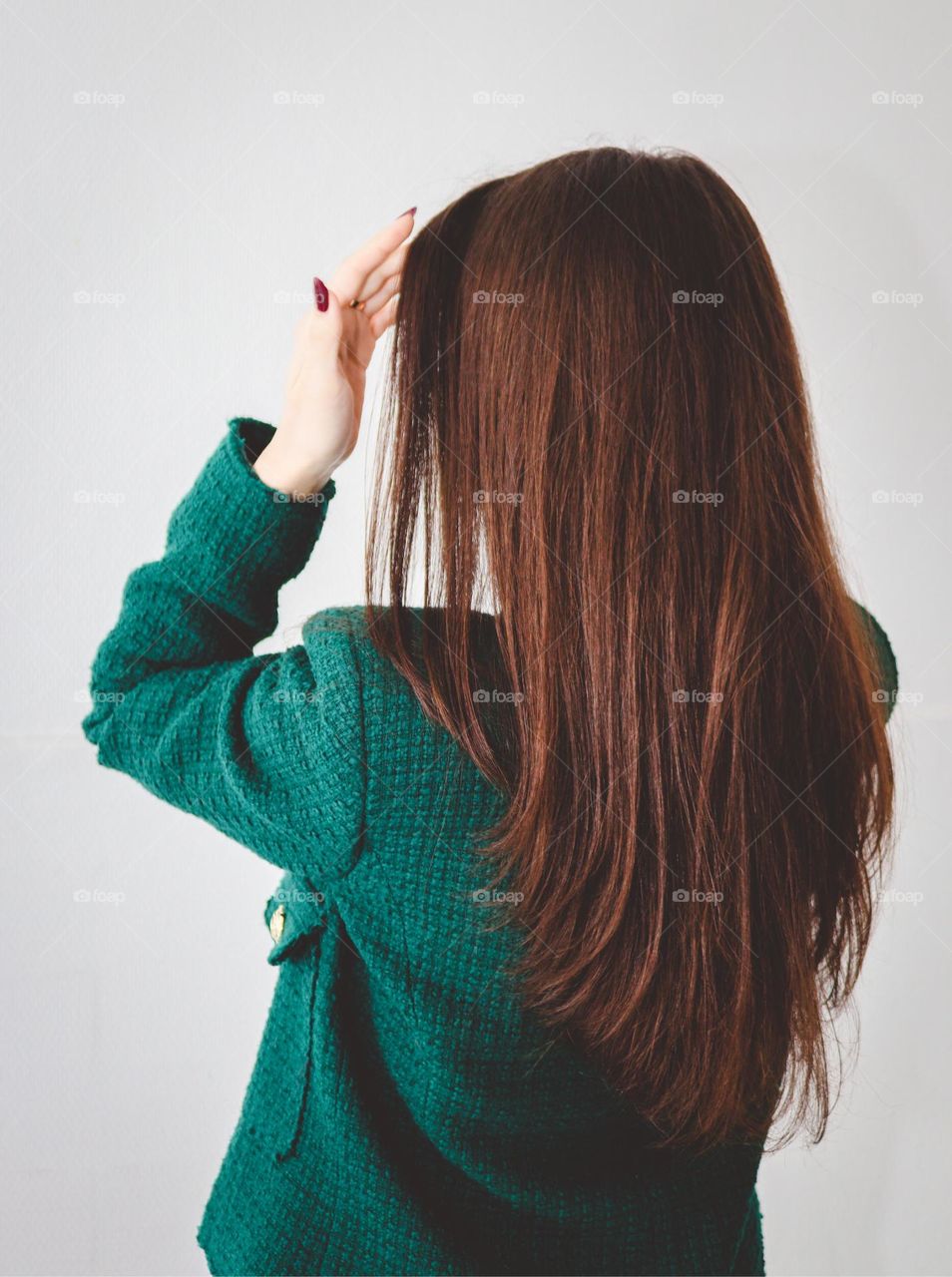 Portrait of beautiful long brown hair with a triangular border in a young Caucasian girl in a green model jacket standing from the back and hand supplying her hair on a gray-white background,side view. The concept of beautiful and healthy hair.
