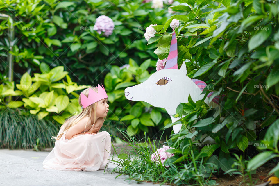 Happy little Caucasian girl with blonde hair and crown standing near unicorn in blooming garden at summer day