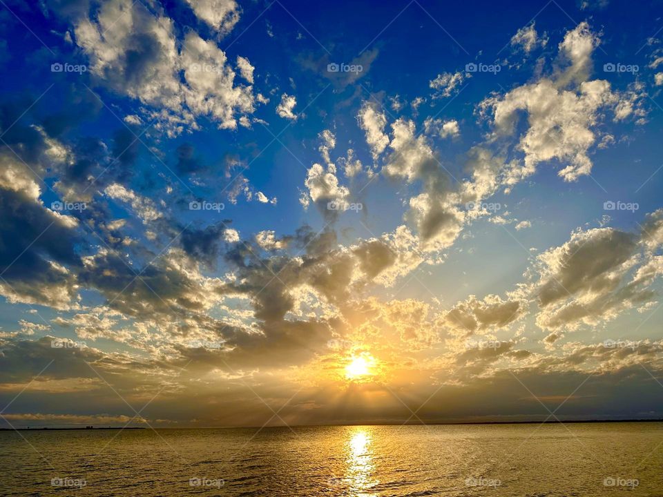 A radiant sunset over the water, with a bright yellow sun near the horizon and explosive pillow clouds scattered across a blue and yellow sky. The sun's reflection is visible on the water's surface.