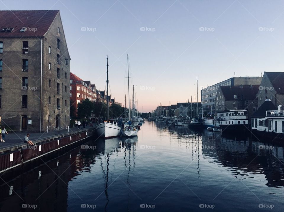 The sun sets over a canal, ships sit in the water and window filled buildings are on either side. 