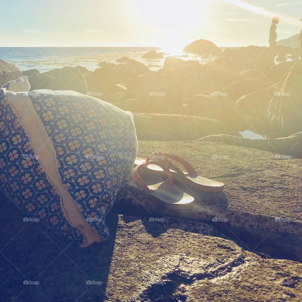 Backlight at the beach focus on flip flops and handbag on the ground and a person by the horizon 