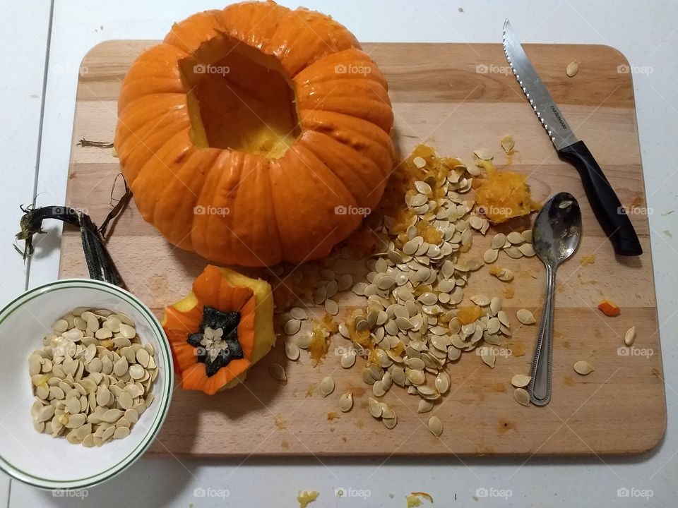 large pumpkin on wooden cutting board being seeded for cooking with removed seeds in bowl, knife and spoon
