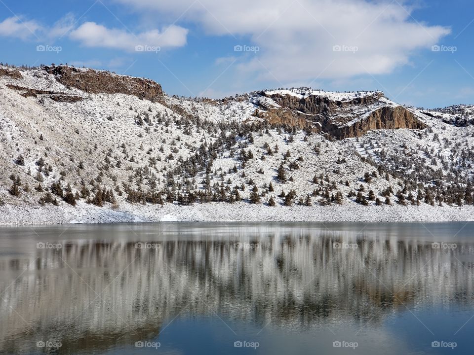 Snow covered hills, trees, clouds, and a bright blue sky reflect in the partially iced over Prineville Reservoir on a sunny winter day in Central Oregon. 