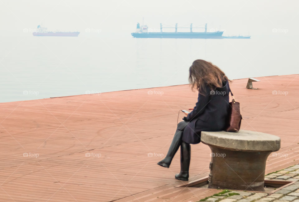 Young Woman Sitting Alone At The Dock Using Her Smartphone And Listening Music
