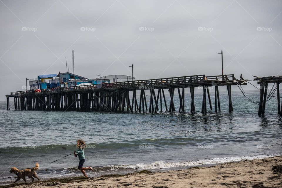 Young girl walking her dog on the beach with the ocean waves on a beautiful day in Capitola California with the wharf in the background 