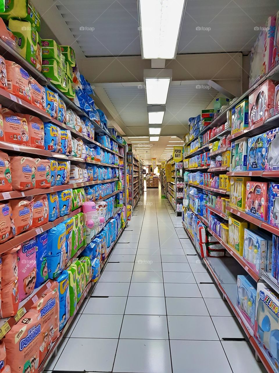 Storefront, baby clothing needs, baby items displayed on opposite shelves, baby diapers, baby clothes, baby pants, baby towels, baby pacifiers, baby hats, racks made of painted steel and labeled product prices