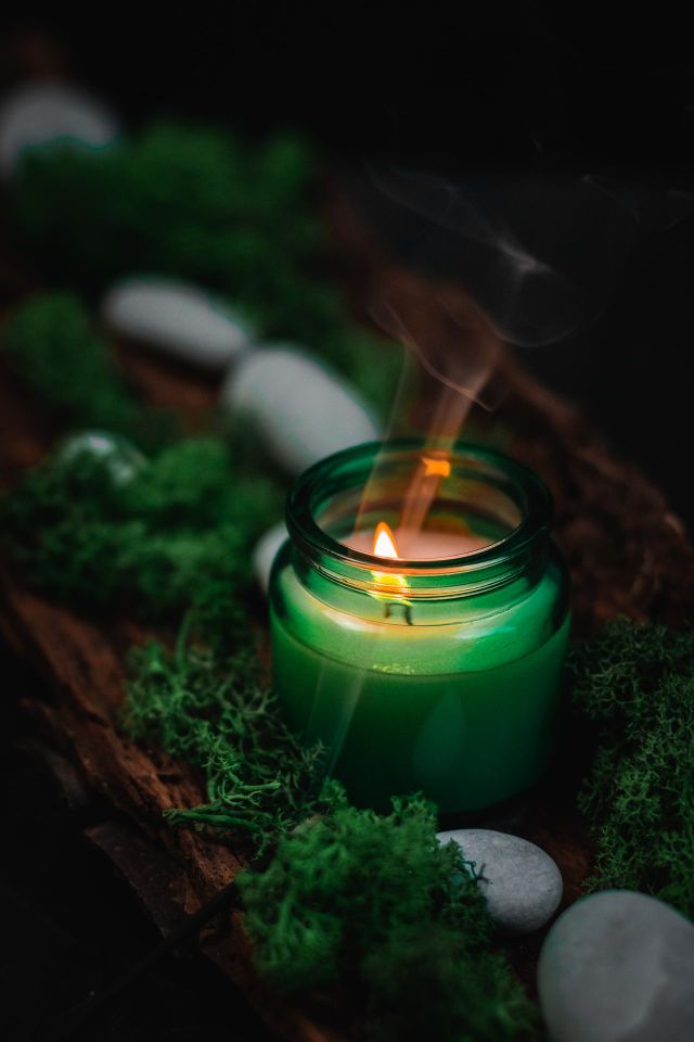 Beautiful view of a candle in a green glass jar standing in a natural tree bark with gray stones, moss and flowing smoke, close-up side view.