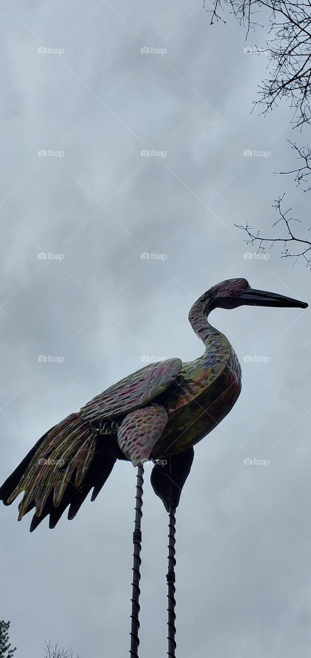 qLarge Heron or Crane standing against a gray overcast cloudy sky. This is a metal sculpture & is a deterrent to scare off other herons.