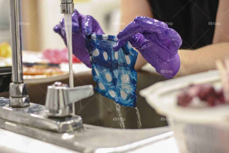 Gloved hands rinse a hand dyed piece of fabric under running water 