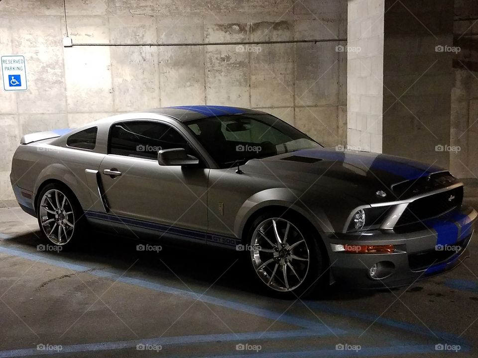 A silver Shelby mustang with blue racing stripes takes up a handicap spot in a garage.