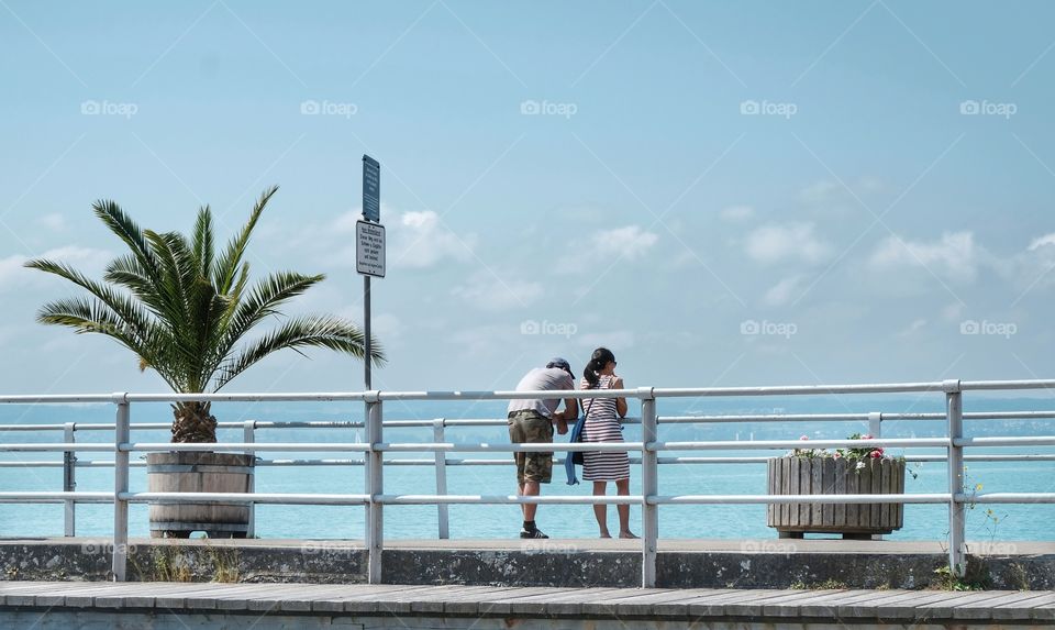 Exotic scenery with palm tree and two persons on a pier looking towards the blue sea. 