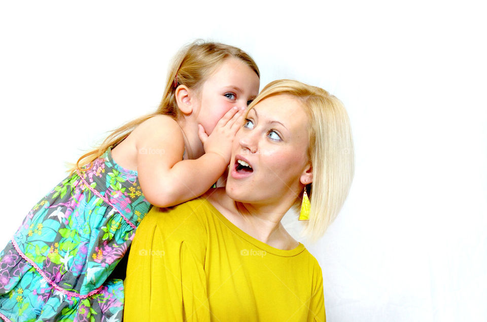 A YOUNG MOTHER LOOKS SURPRISED AS HER DAUGHTER WHISPERS IN HER EAR.