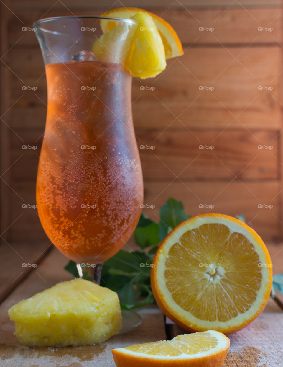 orange non-alcoholic tropical drink in an hourglass shaped glass with fresh pineapple and orange wedges on a wood table with a wood background