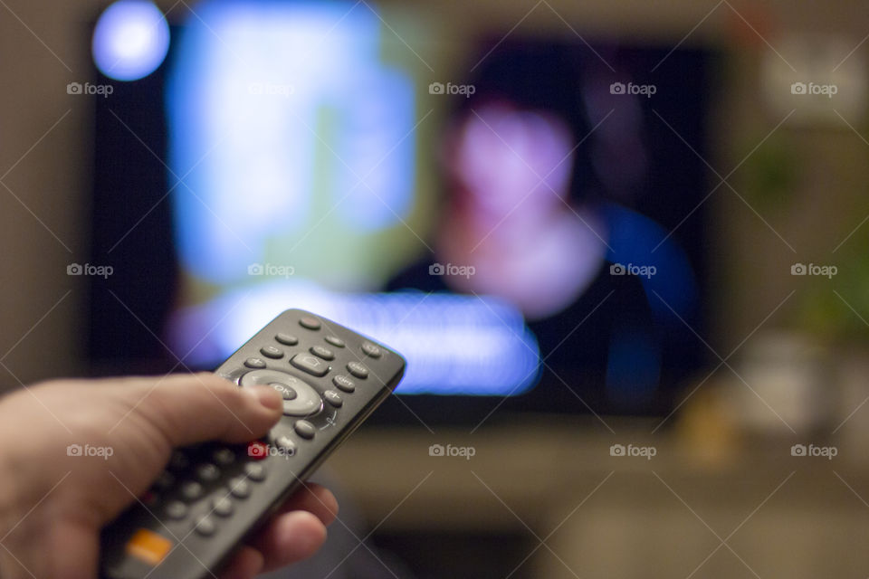 A portrait of a remote control in a hand pointing at a blurry television playing a television show.
