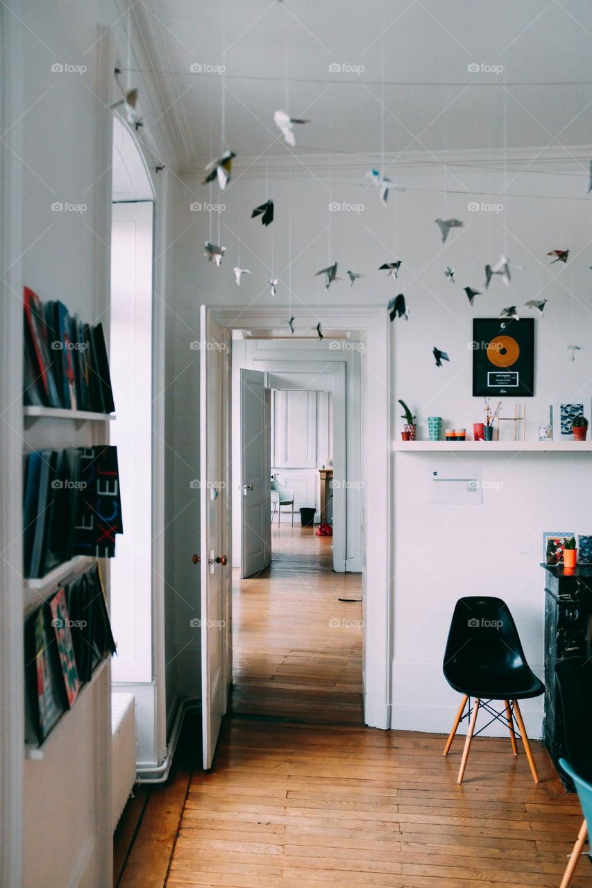 "Origami cranes are hanging from the ceiling of the white room.
In the background, a door is open and you can see inside the apartment."