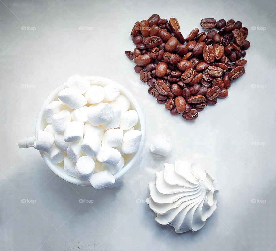 White colour.  On a white background stands a white cup with a white marshmallow, a white marshmallow in the shape of a seashell and brown coffee beans in the shape of a heart.  View from above